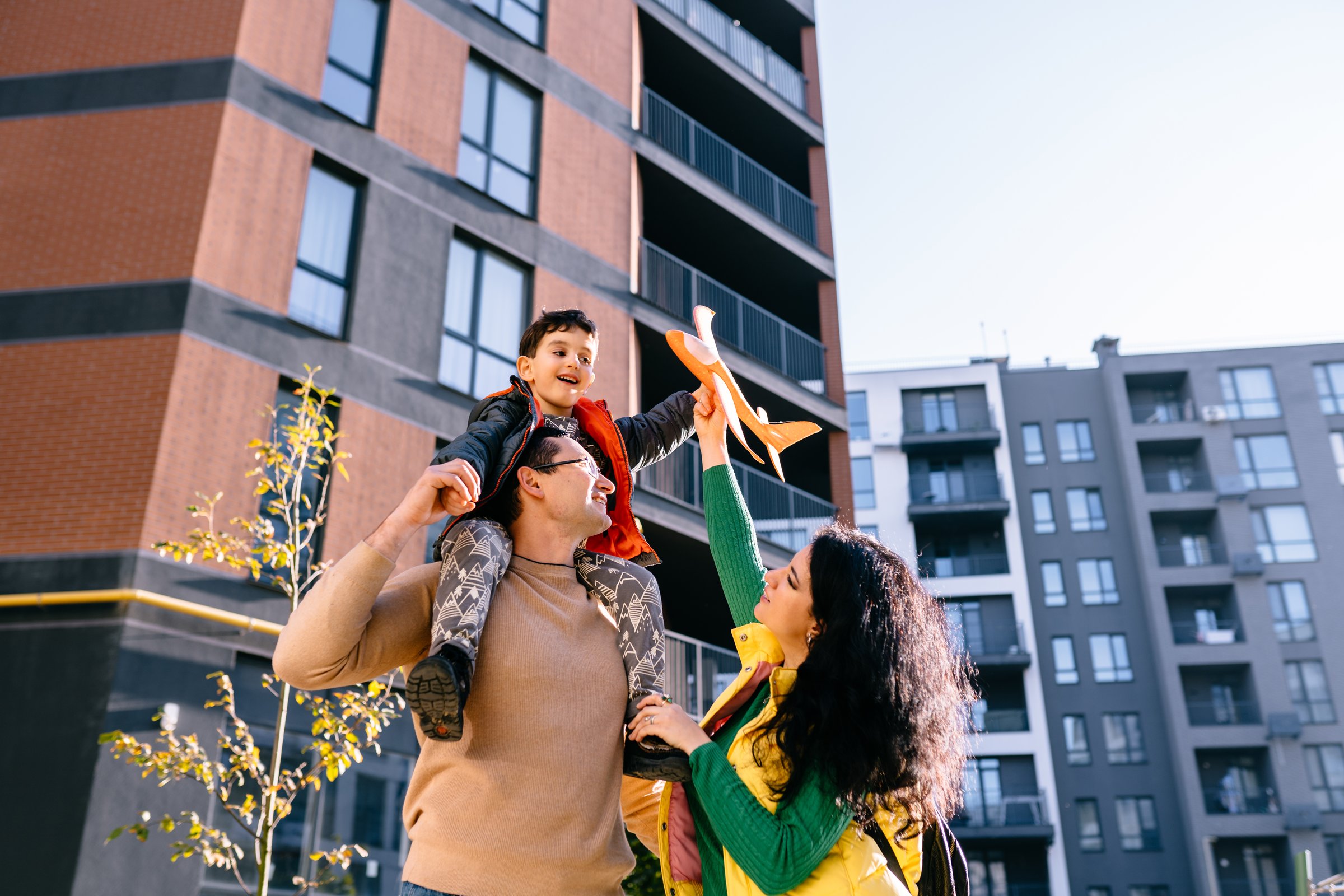 Father and mother, son on father's shoulder smiling in colorful casual outfit holding vibrant orange toy plane near modern apartments. Parents with child play together.