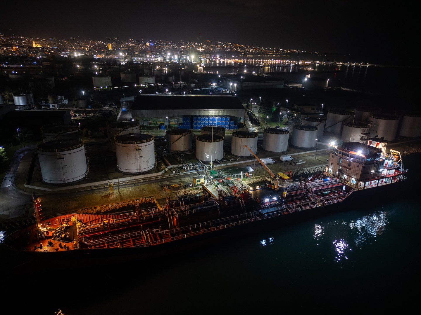 Aerial night photo of an industrial port with an oil tanker moored at the dock, illuminated storage tanks and refinery infrastructure along the waterfront.