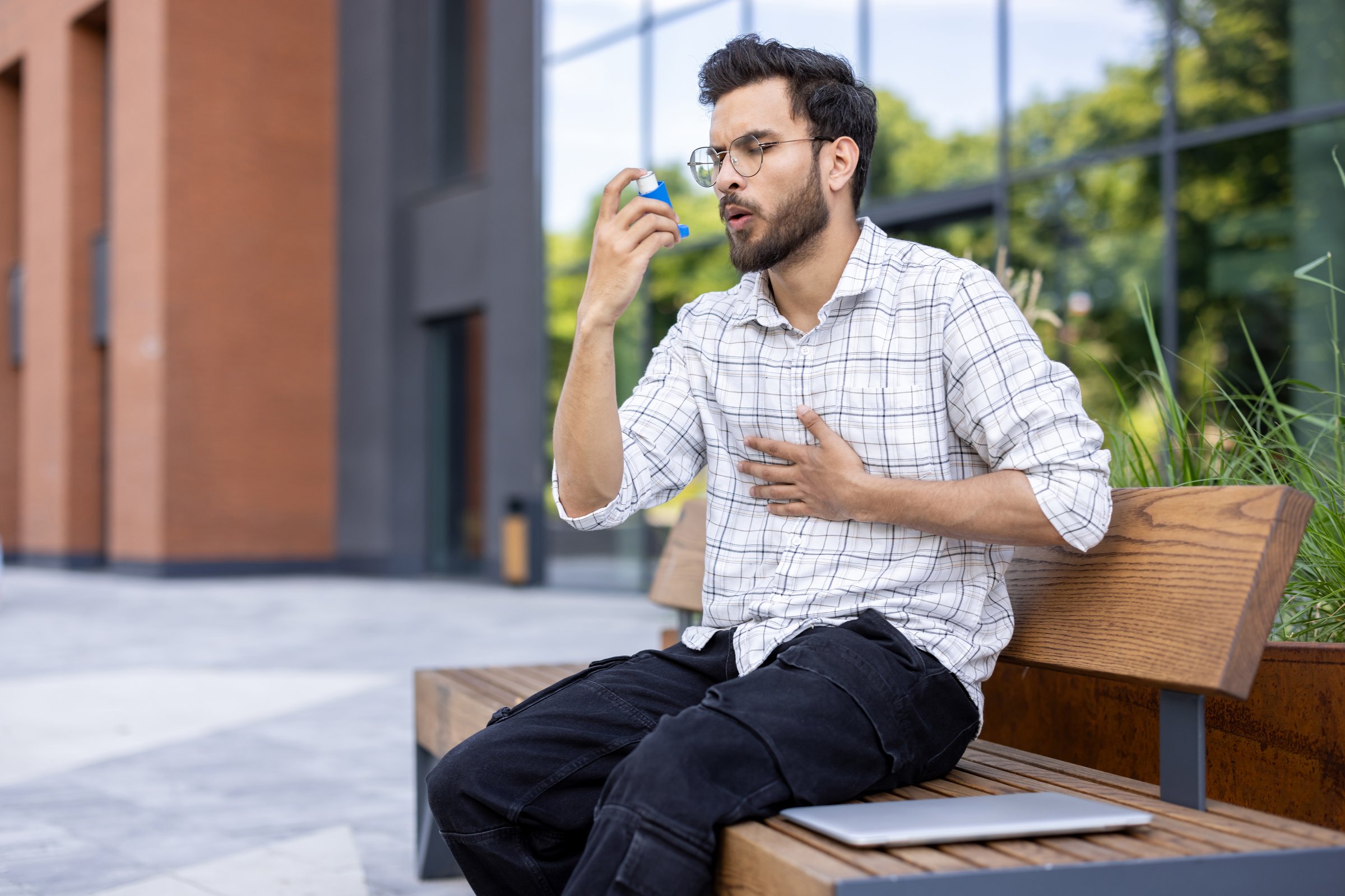 A young Indian man is sitting outside on a bench, suffering from asthma and using an inhaler.