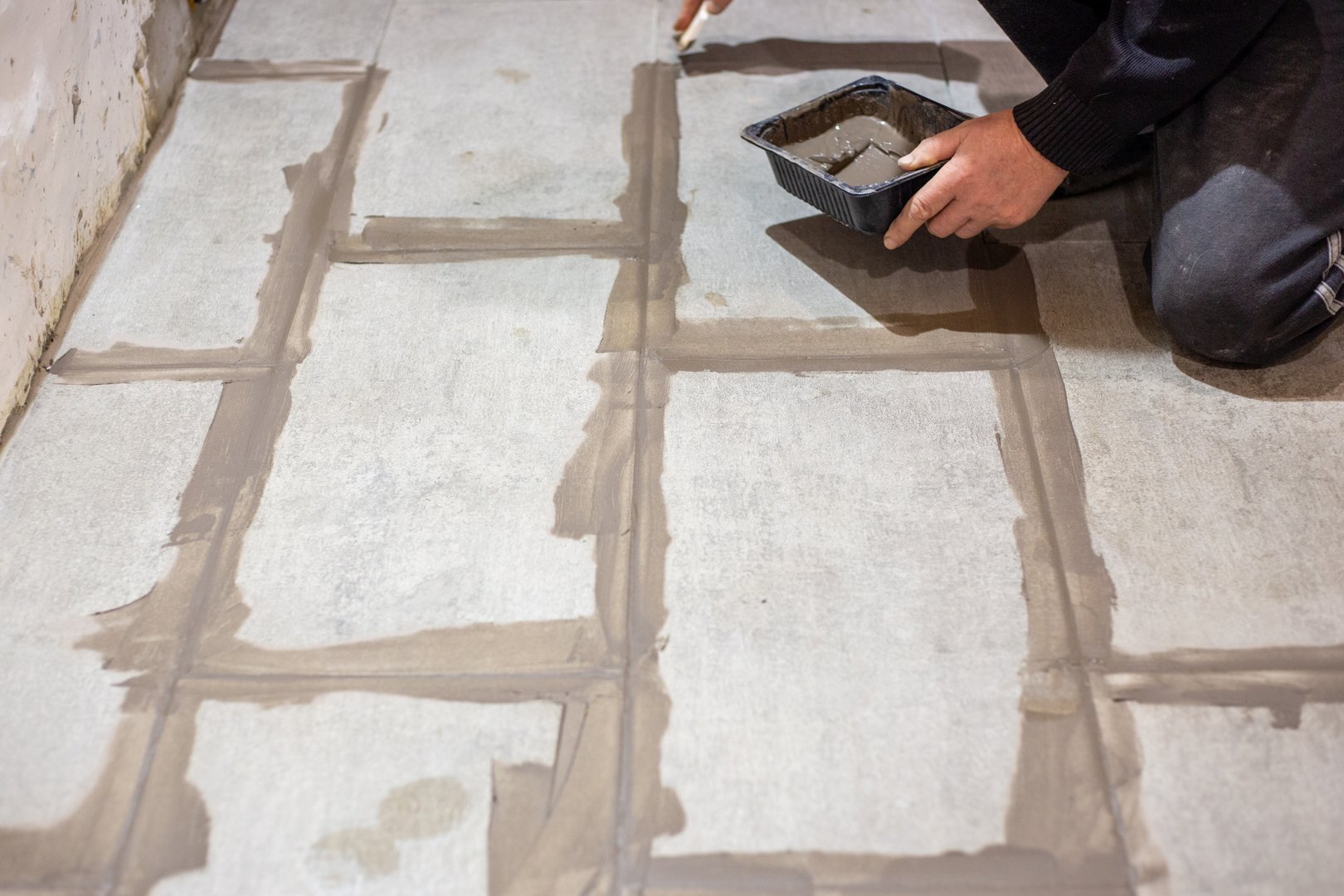 A worker applies grout between newly laid large floor tiles using a small container for the material.