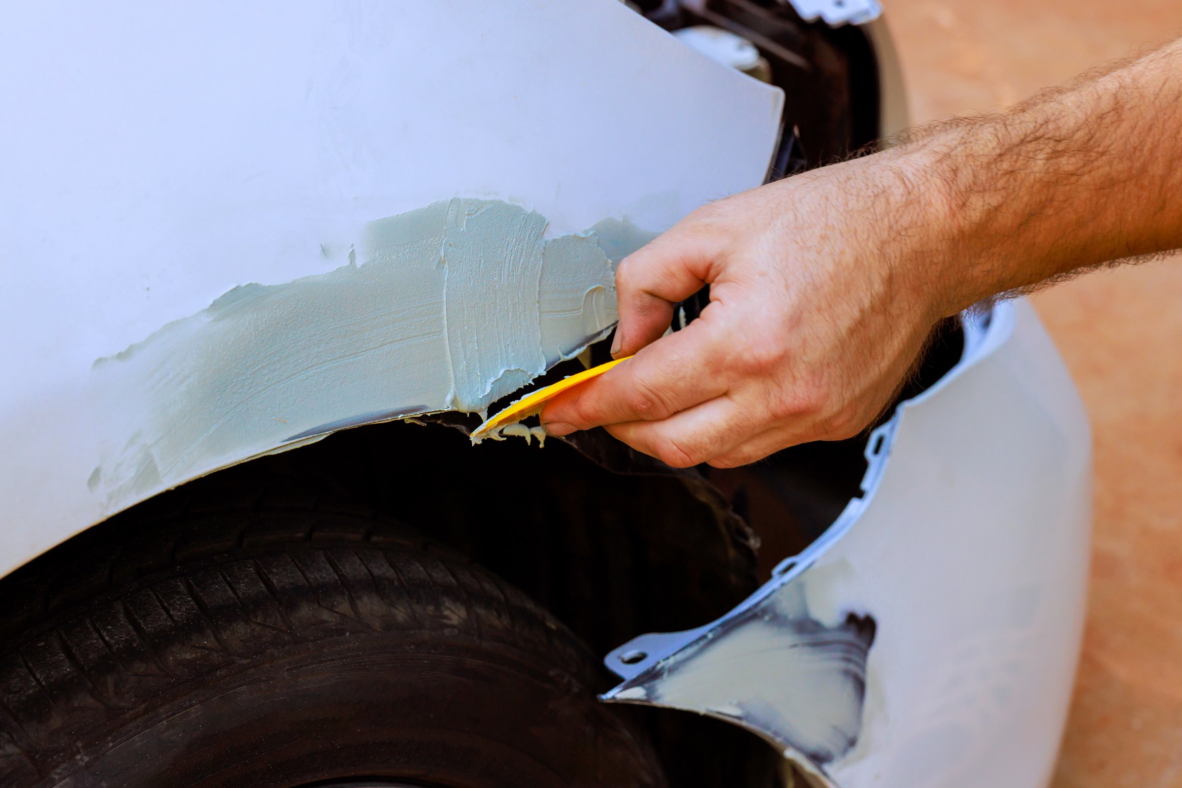 Worker uses tool to smooth body filler over car dent in repair shop, vehicle restoration work.