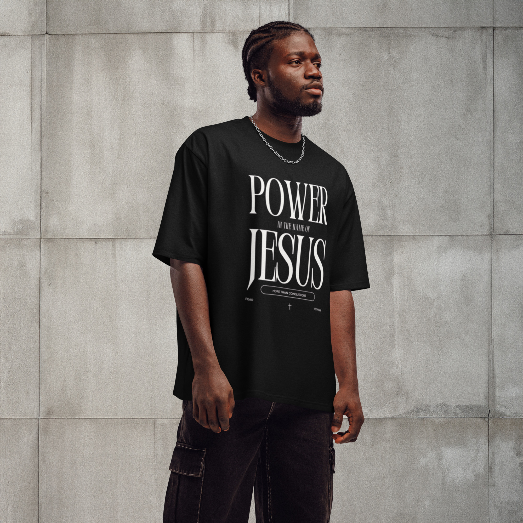 Man in a black "POWER of the NAME of JESUS" T-shirt stands against concrete wall, casually posing with hands at sides.
