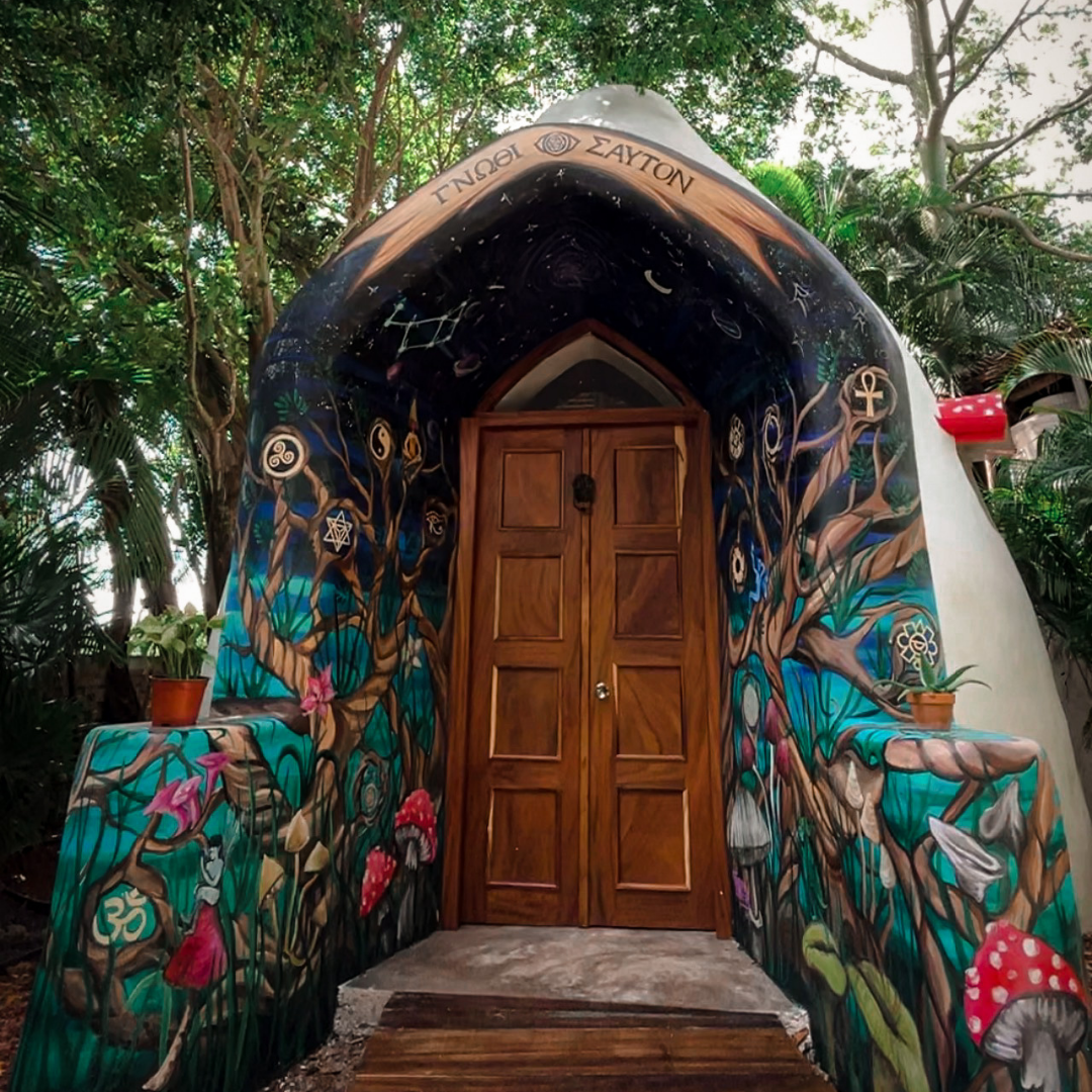 Whimsical doorway with colorful mushroom and nature mural, surrounded by lush greenery; archway reads "T. NGOI & ZAYTON."