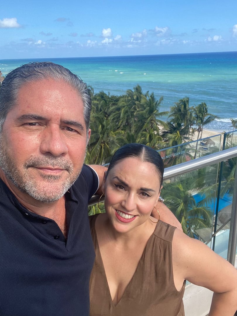A man and woman taking a selfie on a balcony with palm trees and the ocean in the background.