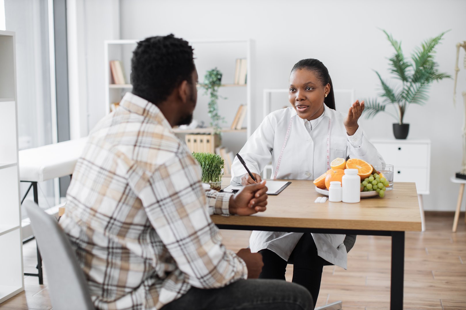 Focused multiethnic woman in doctor's coat talking to middle-aged man in consulting room of hospital. Professional expert in nutrition providing personalized guidance to male client on food habits.