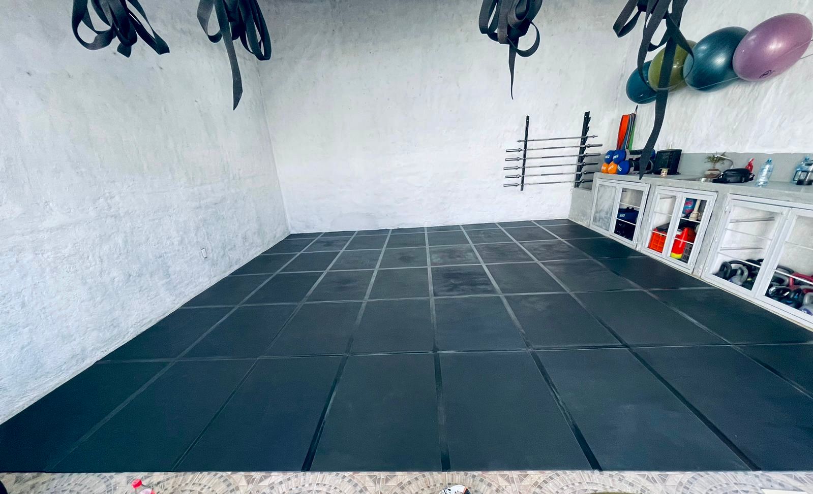 A home gym with black exercise mats, wall-mounted straps, colorful exercise balls, and various gym equipment on shelves.