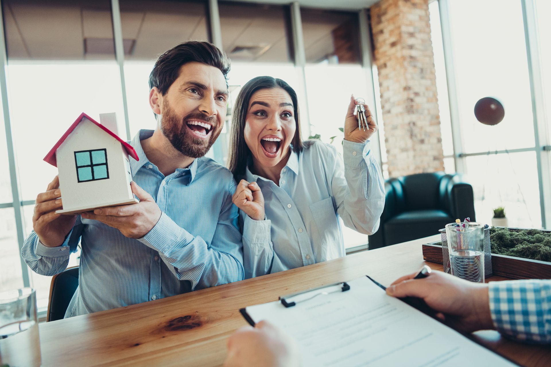 A happy couple sitting at a desk, holding keys and a house model, symbolizing success in real estate or property ownership.