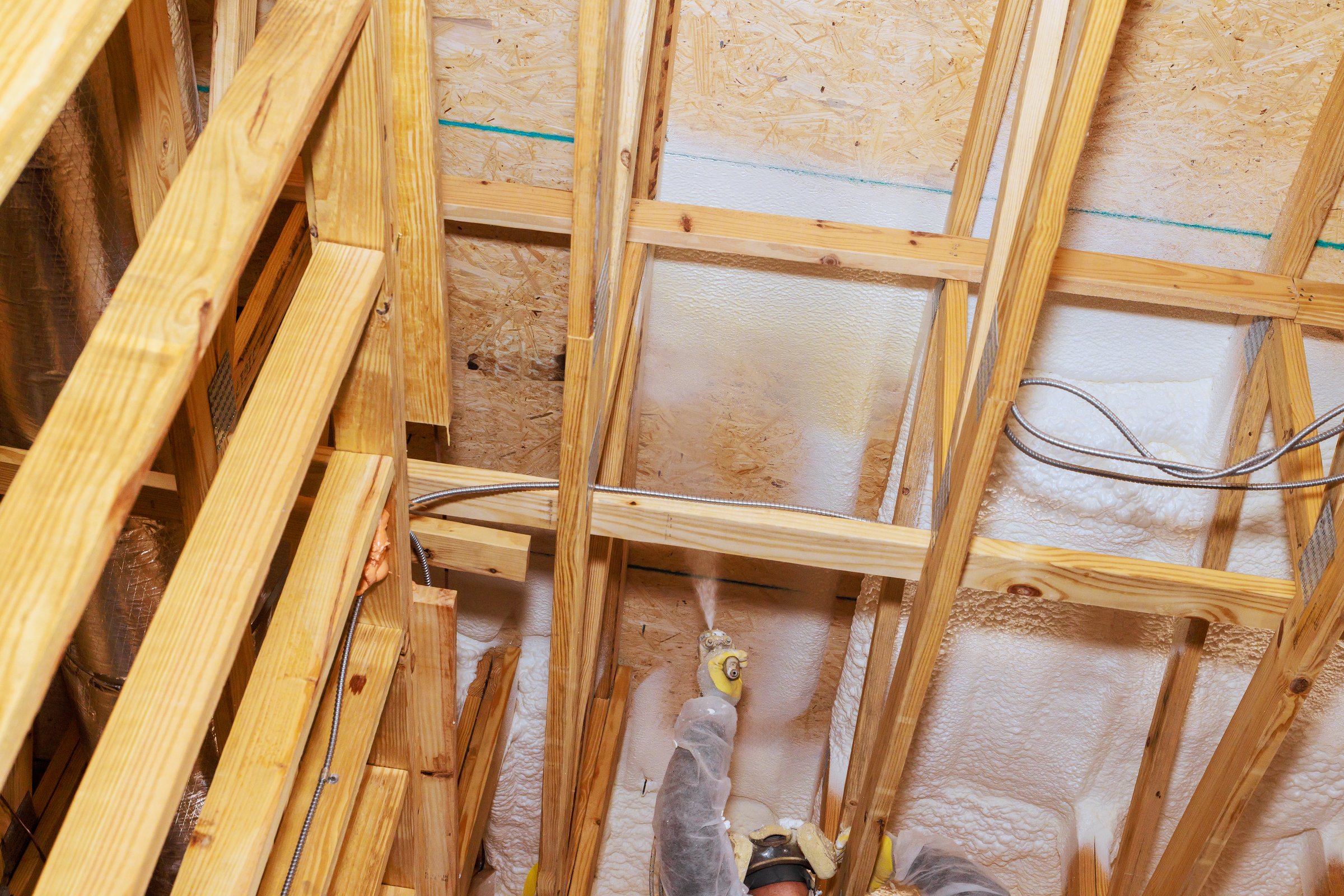 Skilled worker after install wooden beams, applies foam insulation material on ceiling framework in new house