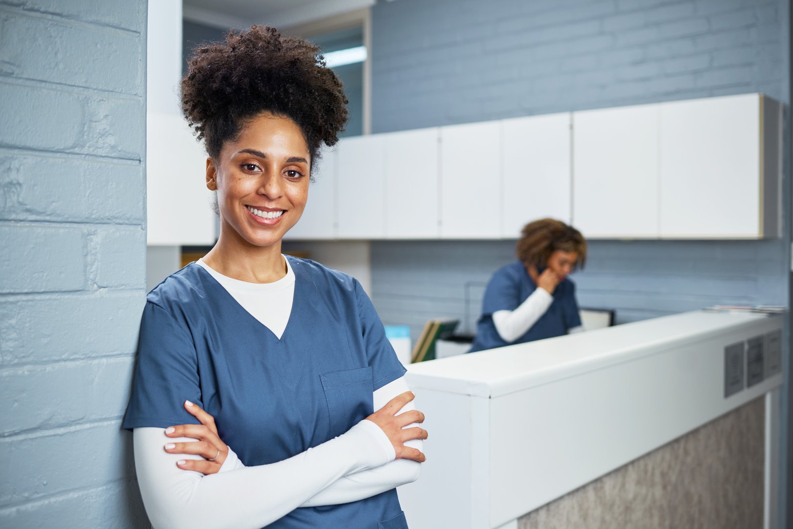 A confident health professional wearing scrubs is standing with arms crossed in a bright and clean dental office reception area. She conveys trust, professionalism, and approachability in a medical environment.