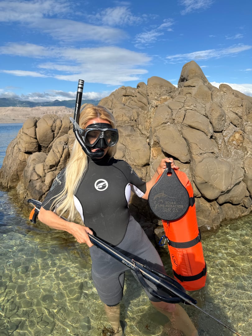 Woman in snorkeling gear stands in clear water holding an orange float and snorkel mask, rocky background and blue sky.