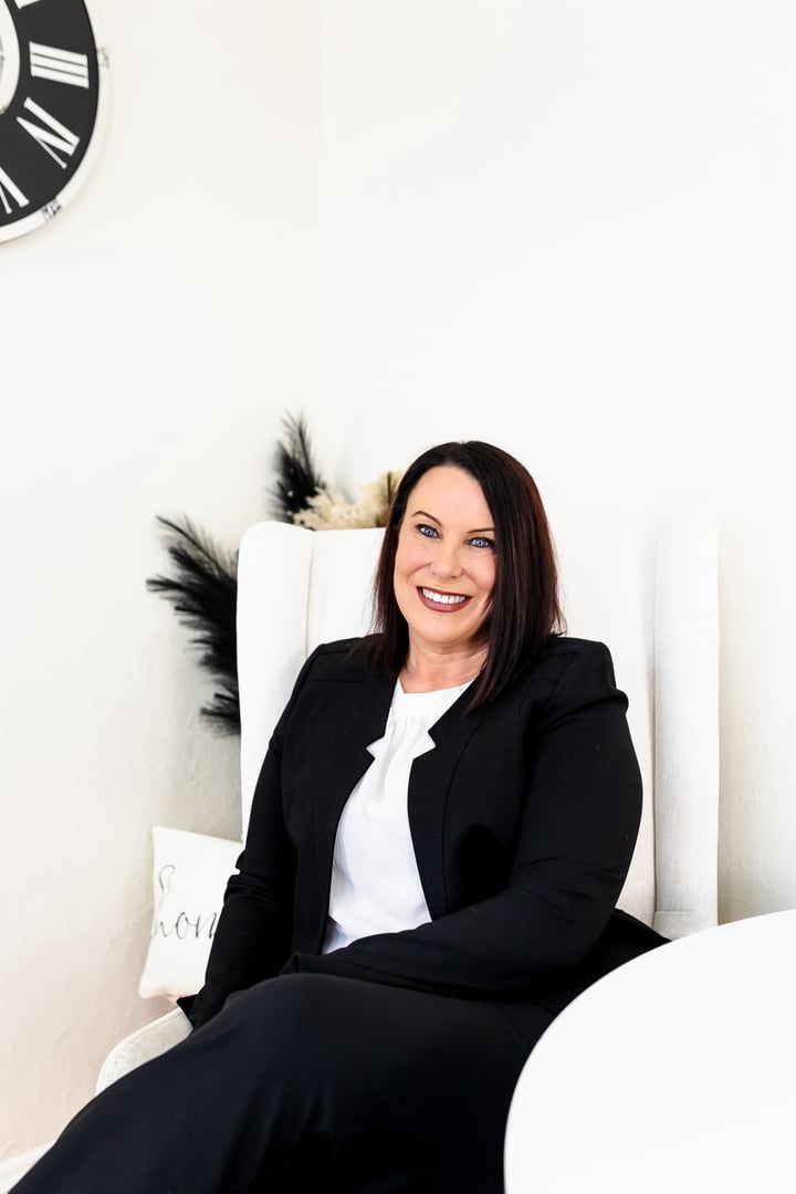 Smiling woman in a black suit sits in a white chair against a light background, with a clock partially visible on the wall.