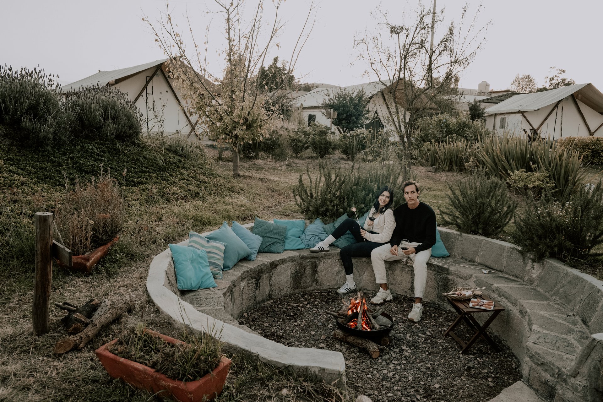 A couple sits by a fire pit on a stone bench, surrounded by cushions, with tents and greenery in the background.
