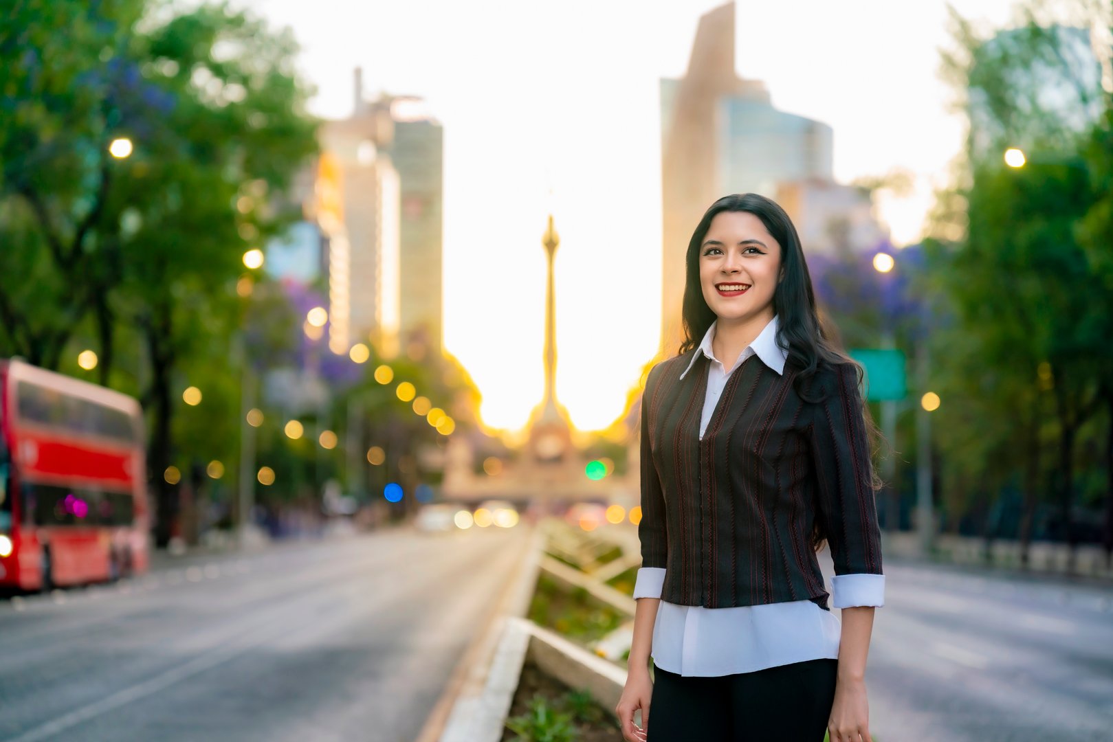 Young latina woman with long hair and business casual outfit stands confidently on Reforma avenue during sunset in Mexico City.