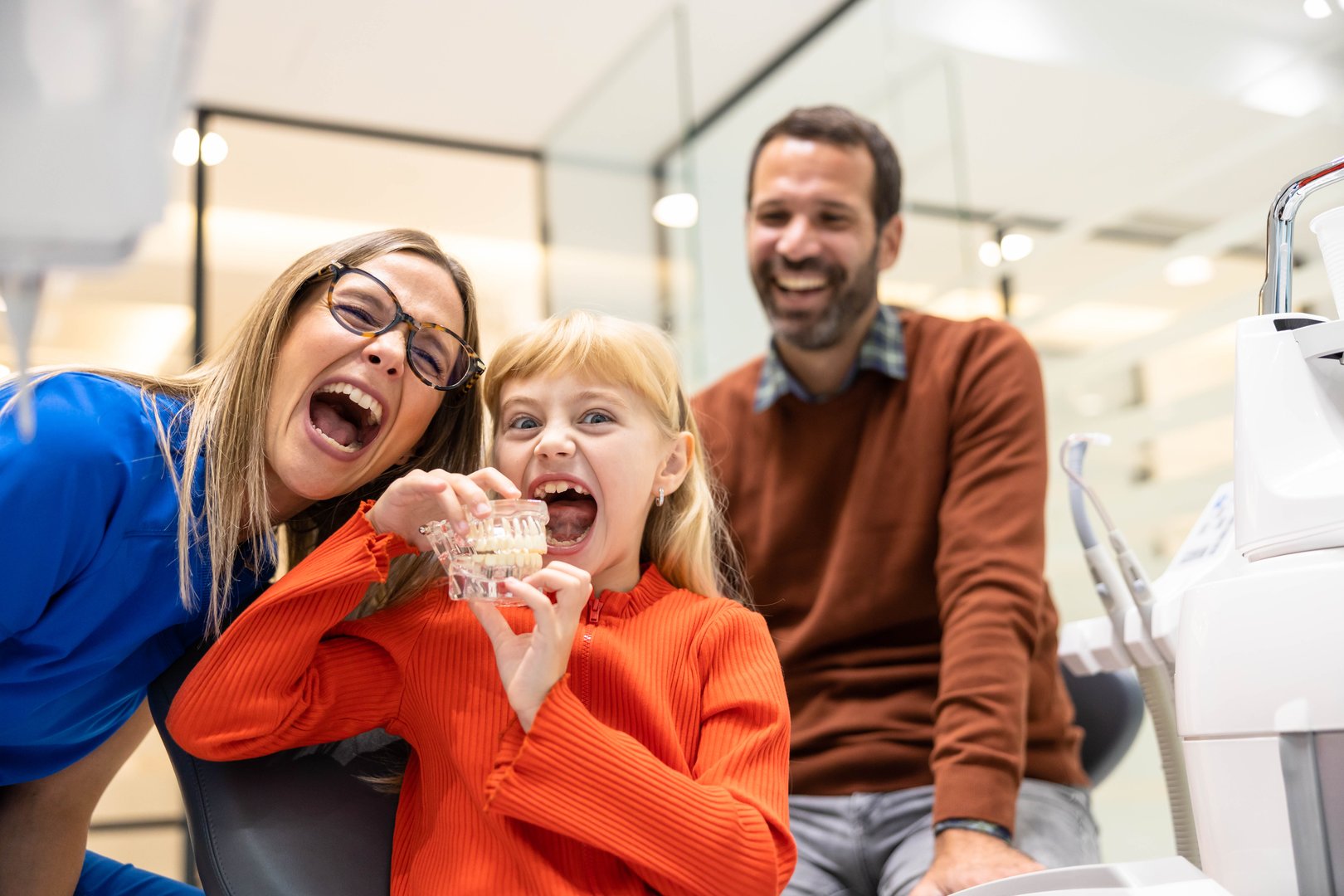 A child shares a joyful moment with a dentist using a dental model, promoting comfort and positivity in the dental clinic environment, father watching.