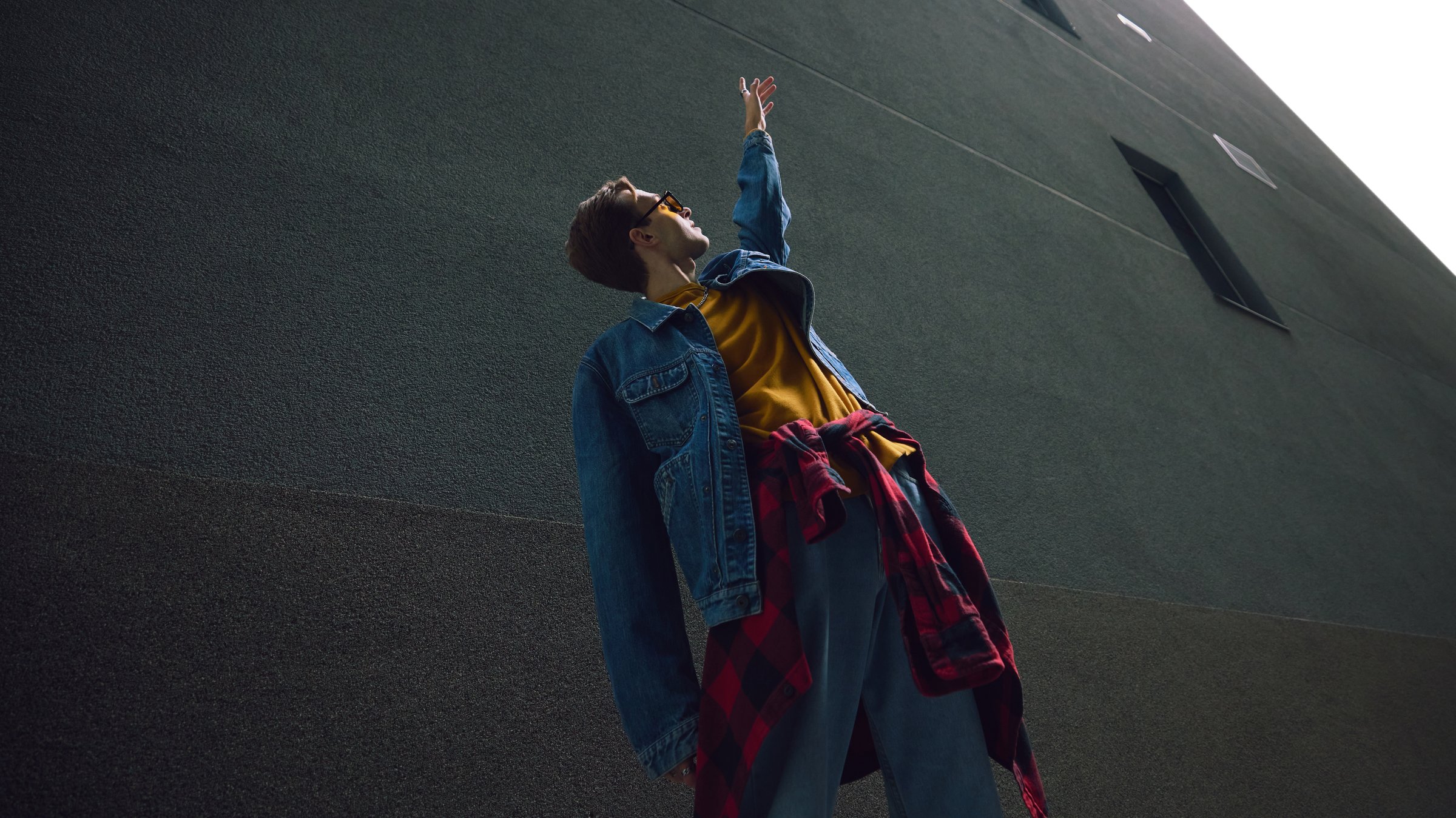 Man reaching upward in denim and yellow outfit near tall building. Concept of motivation in lifestyle, education promotion, creative learning, urban communication, and visual identity.