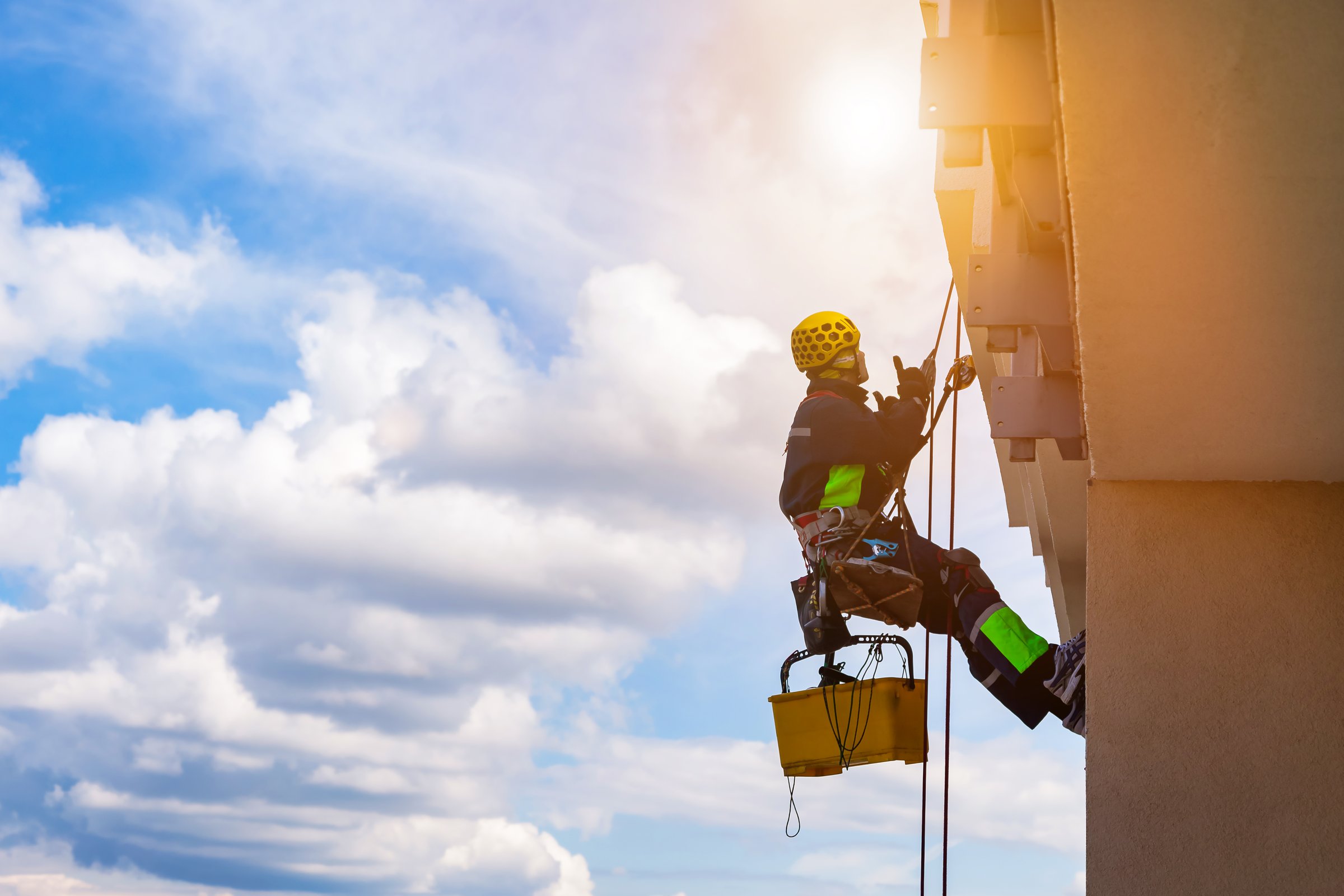 Industrial mountaineering worker washing exterior facade glazing hangs over residential facade building while at blue sky background. Rope access laborer hangs on wall of house. Copy space