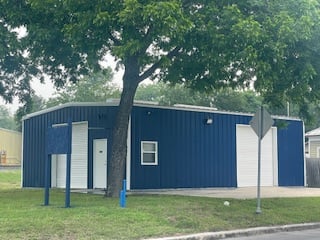 Blue metal building with two white garage doors, a tree in front, and a blank sign on grass. Overcast day.