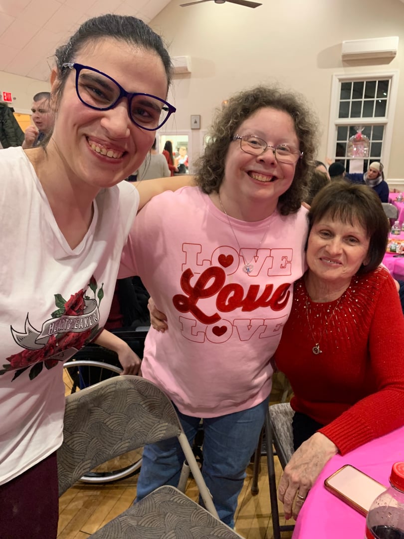 Three women smiling at an indoor event, one wearing a "Love" shirt, another in red, seated at a table with pink decorations.