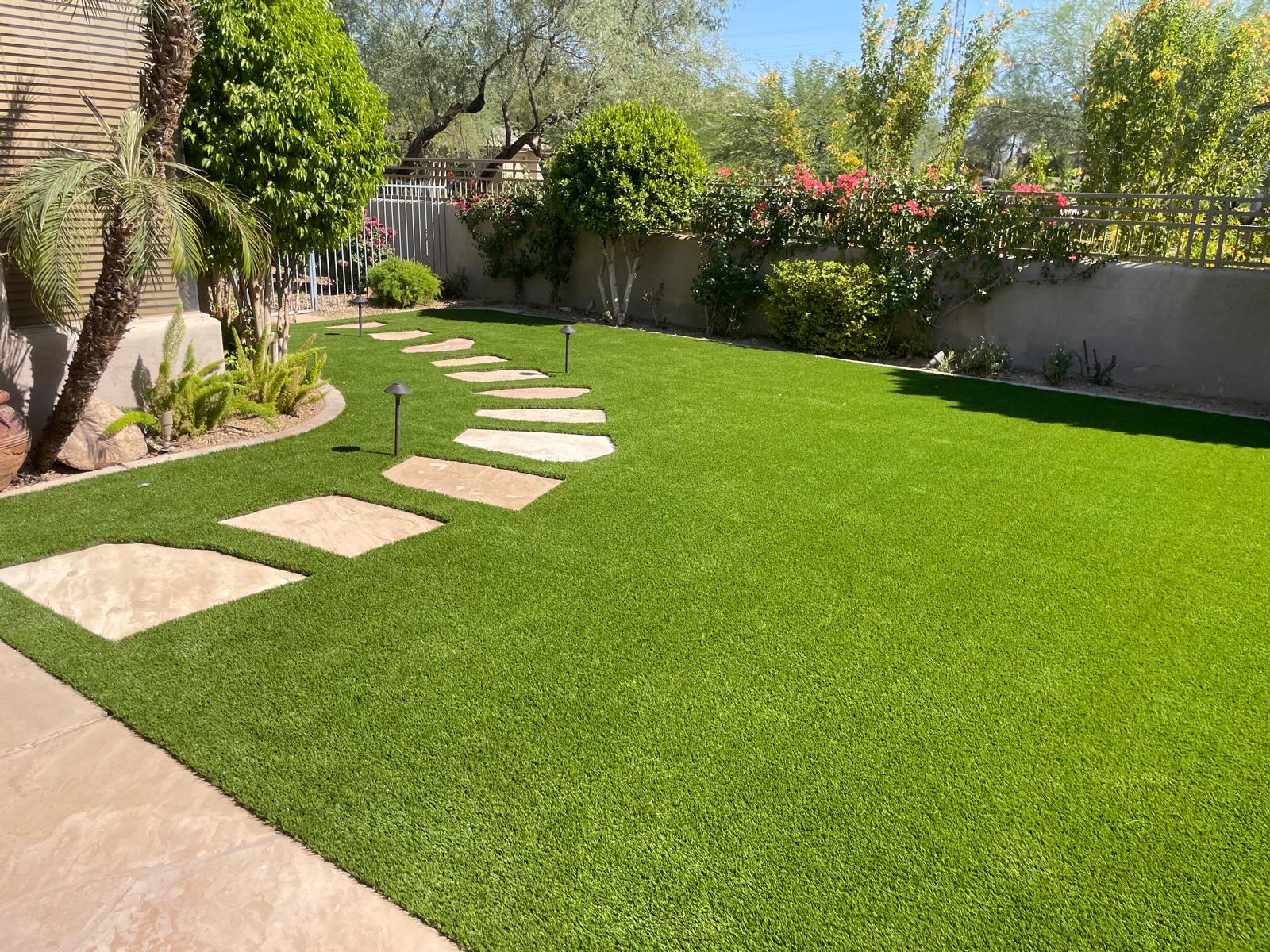 Backyard with artificial grass, stone path, and landscaped plants, bordered by a fence with blooming bushes.