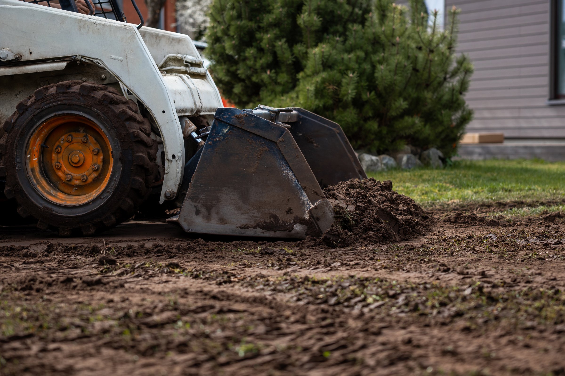 A skid steer loader with orange wheel rims moves soil with its bucket. Nearby are a green bush, a building with horizontal siding, and excavated ground.