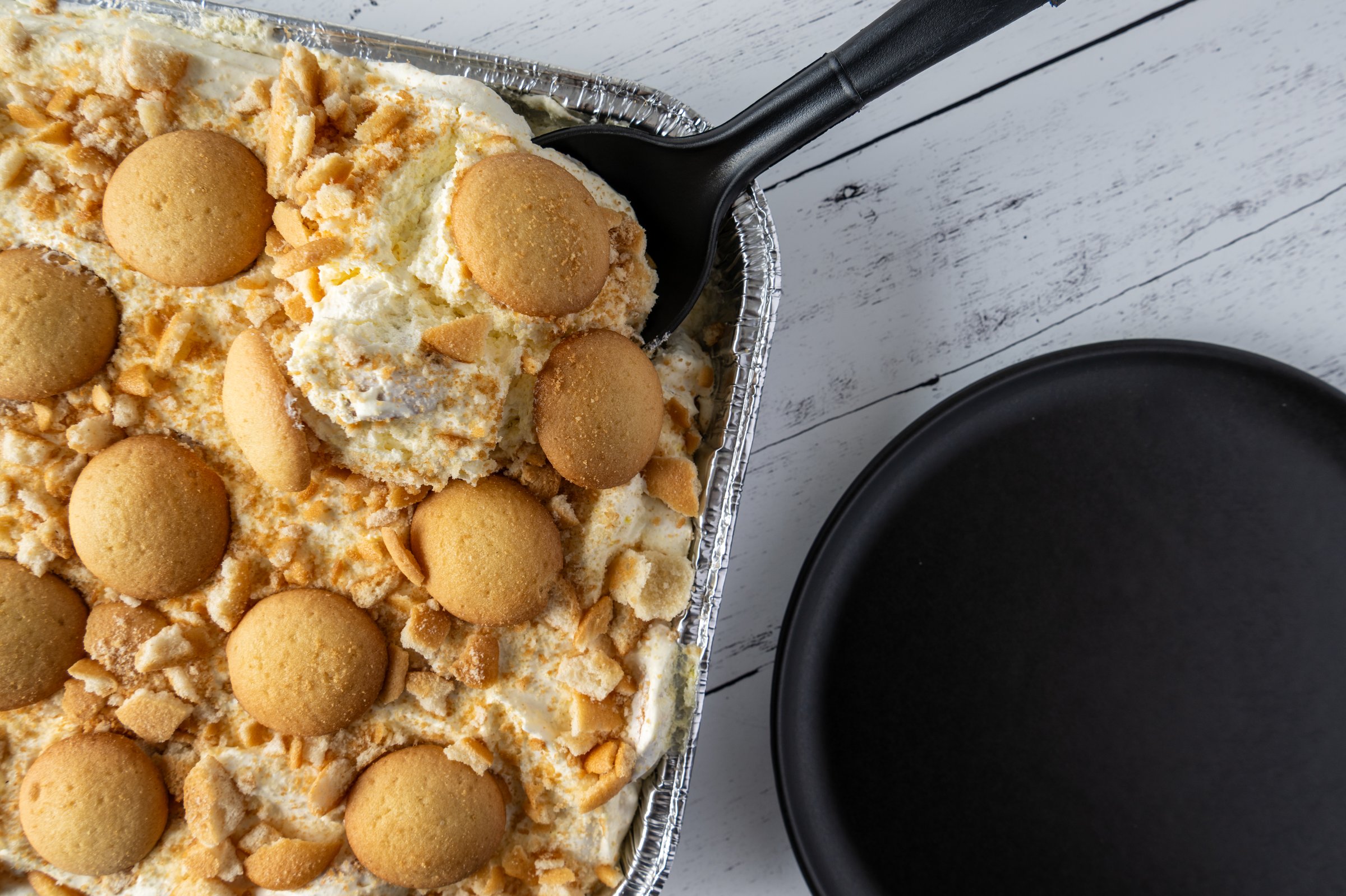 Flat lay of a tray of homemade Banana pudding with vanilla wafer cookies on top with a serving spoon in it and a black plate next to it on top a wood table.