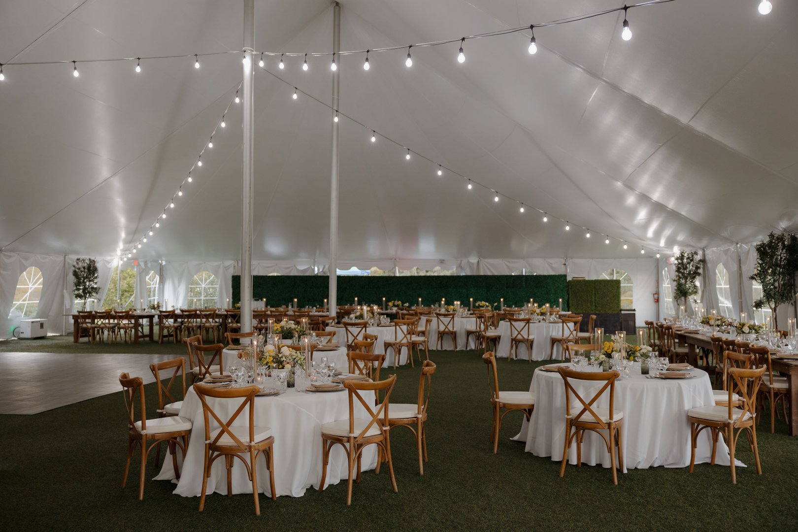 A beautifully decorated wedding reception tent with round tables, wooden chairs, and string lights creating a romantic ambiance.