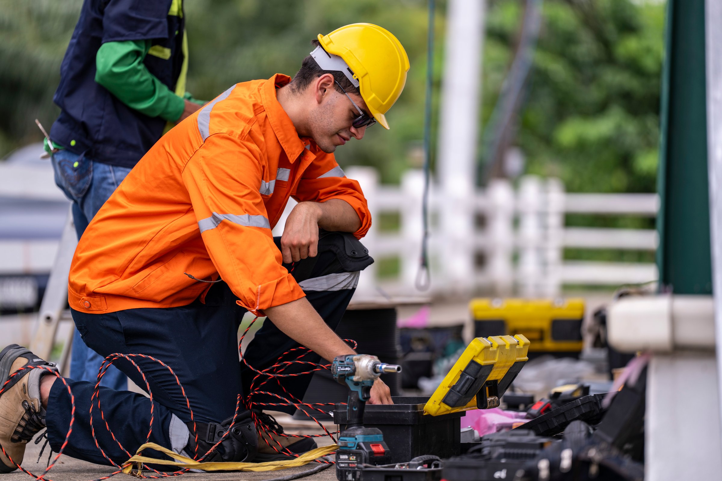 A skilled technician in a bright orange safety jacket and hard hat prepares electrical wiring with precision. Tools and open cases are spread in the background, showcasing a professional workspace.