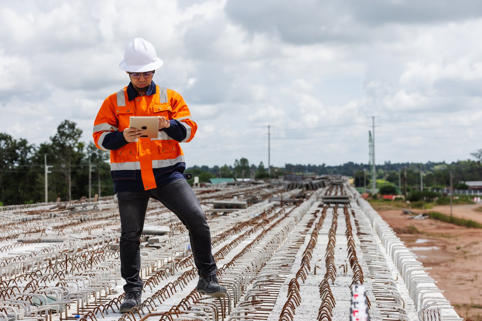 A construction engineer wearing a white helmet and orange safety jacket stands on a bridge under construction, using a digital tablet to inspect steel reinforcement and monitor project progress.