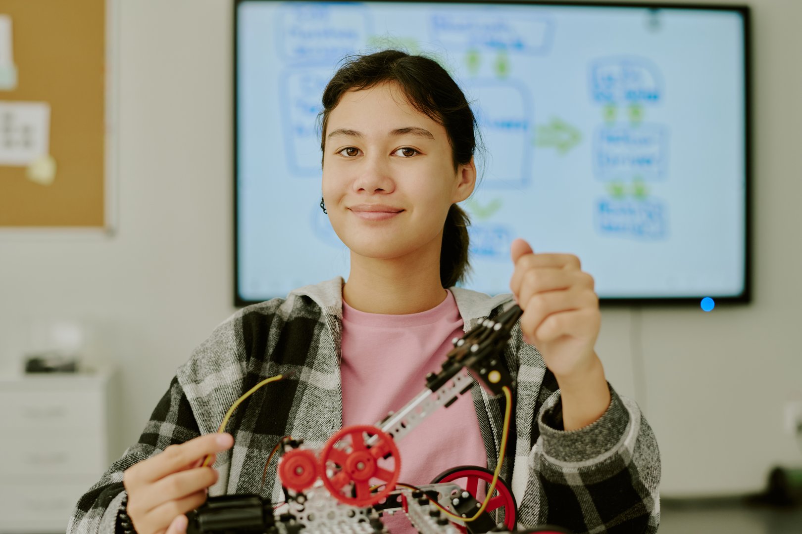 Portrait of smart girl holding wires of robot which on table while posing for photo in classroom