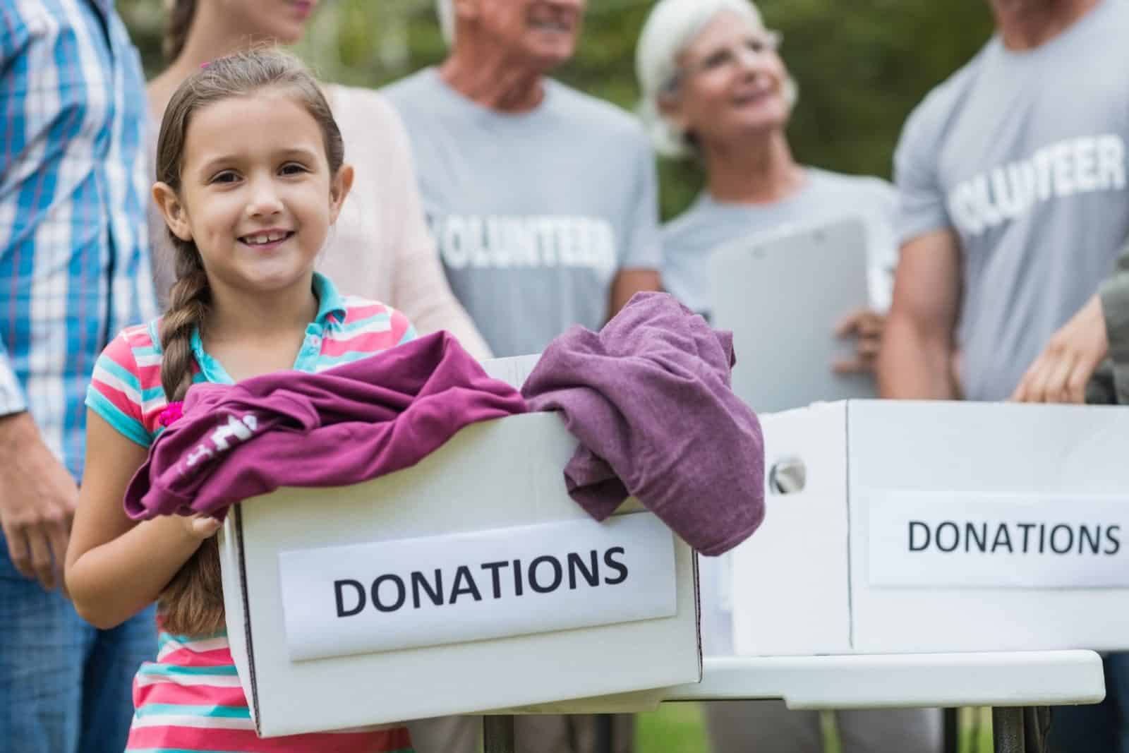 A young girl smiles while holding a box labeled Donations, surrounded by adults wearing volunteer shirts.