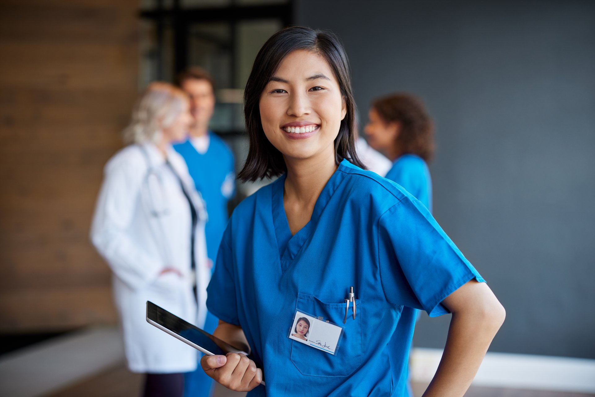 Cheerful asian nurse holding digital tablet with healthcare team in the background and looking at camera. Portrait of confident medical professional in blue scrubs ready to assist. Portrait of female nurse smiling, with healthcare workers standing in background.