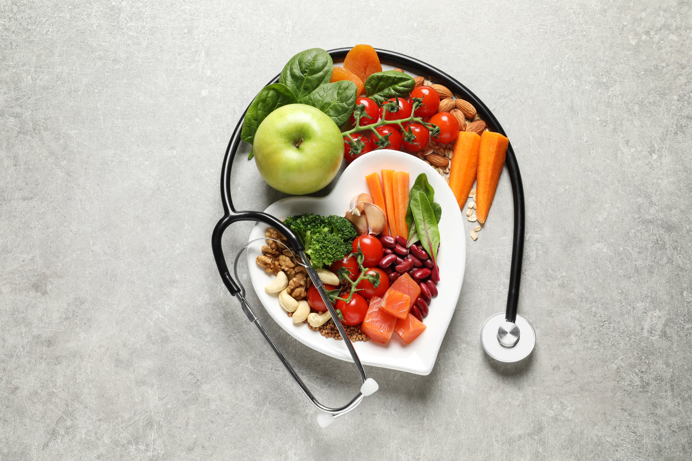 Overhead view of a heart-shaped plate containing a balanced diet rich in protein, fiber, and vitamins, including carrots, broccoli, tomatoes, apple, walnuts, and fish. A doctor's stethoscope is draped around the plate on a textured gray background, symbolizing cardiovascular wellness and dietary health. Perfect for articles on nutrition, cardiology, preventative medicine, and healthy lifestyle promotions.