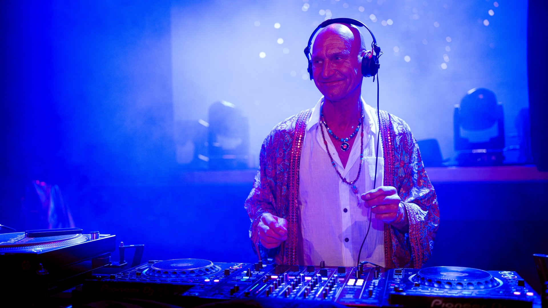 DJ performing on stage with headphones, wearing a patterned jacket in a blue-lit atmosphere.