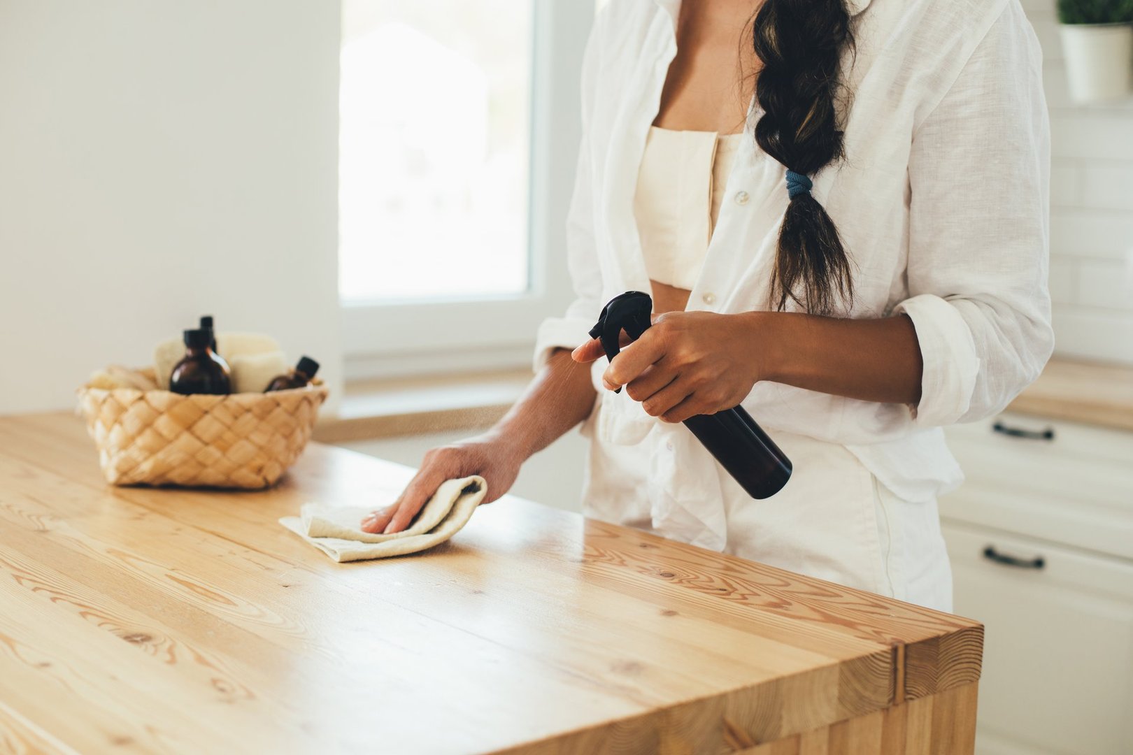 Young woman cleaning wooden table using spray in glass bottle and natural rag in a kitchen.