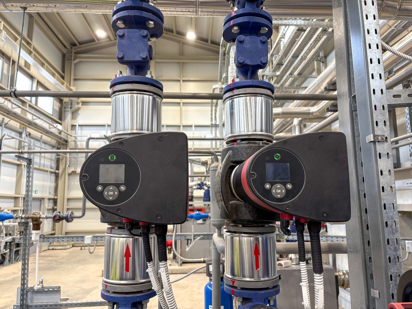 Industrial water pump system inside a modern facility, featuring labeled control panels and stainless steel piping, used for automation, plumbing, or energy systems.