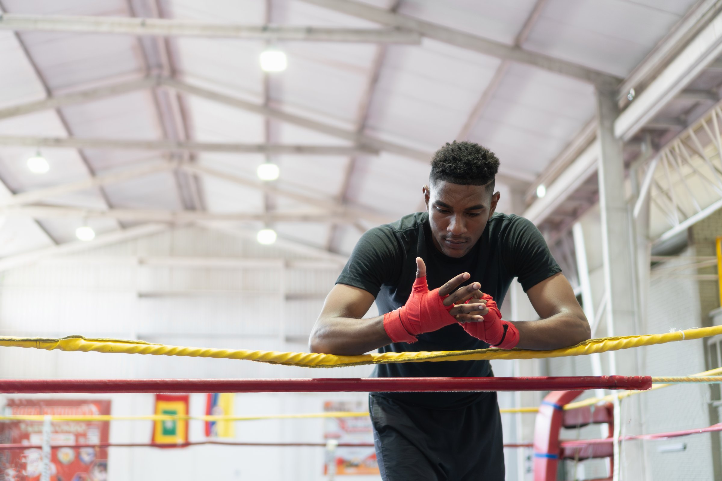 Young adult boxer with hand wraps leaning on the ropes of a boxing ring, looking downwards in thought