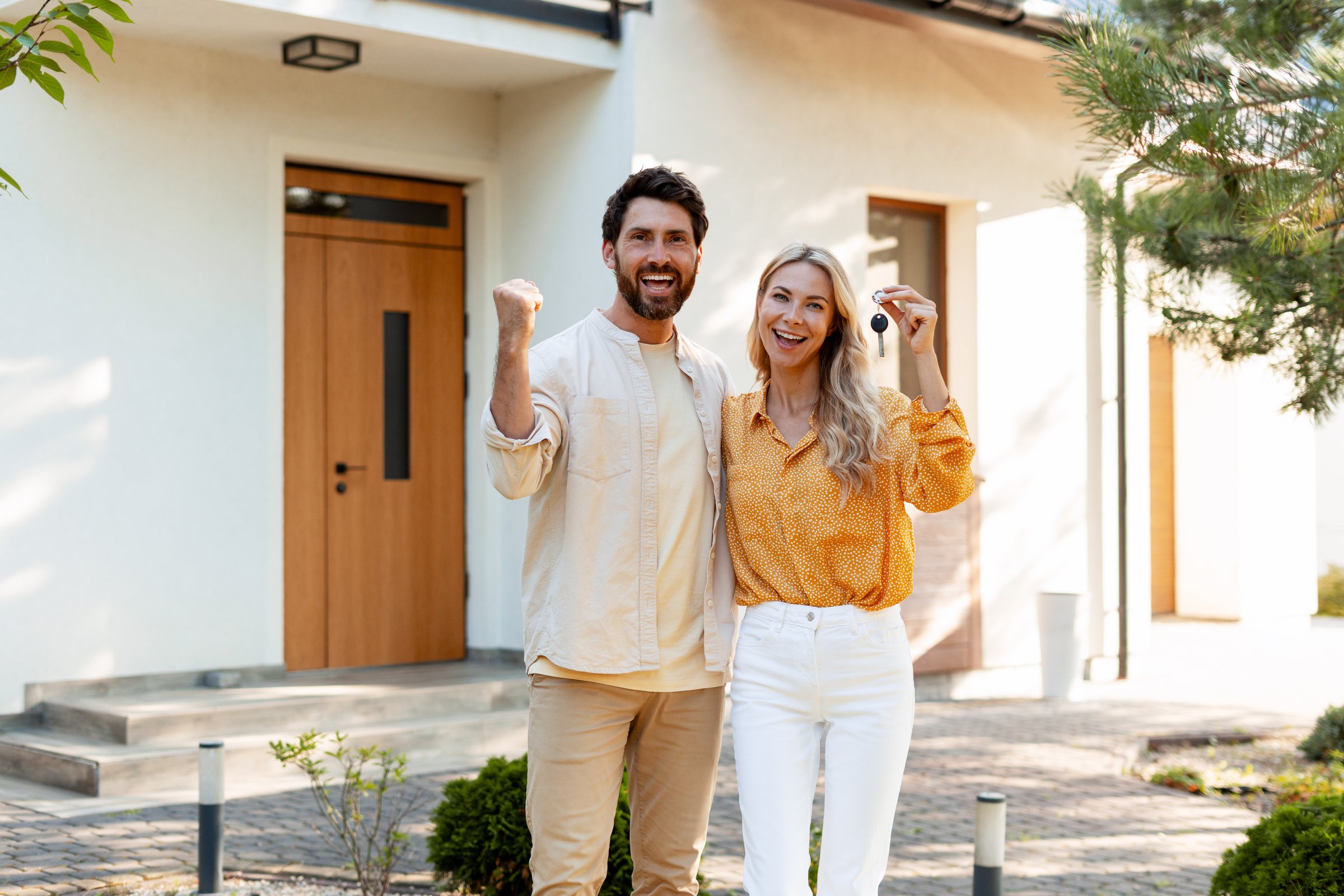 Excited couple celebrating their new home purchase, proudly displaying keys in front of their beautiful property, embodying the joy of homeownership and family togetherness