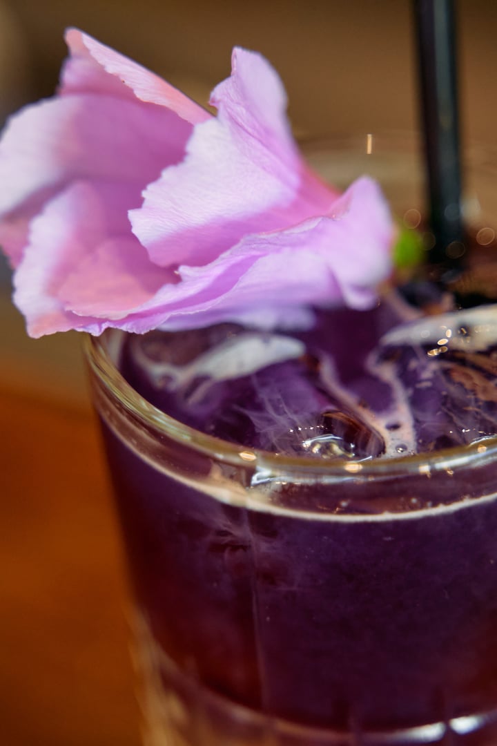 Vibrant purple beverage in a clear glass, adorned with ice and a pink floral garnish. aesthetic and refreshing choice for a sophisticated drink.
