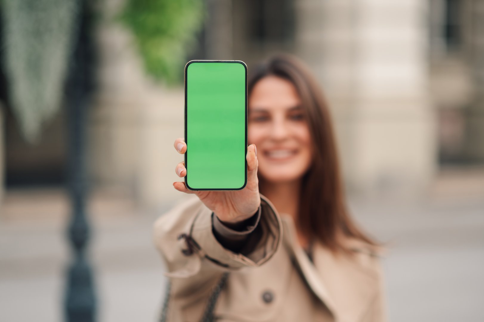 Young woman happily smiles, holding a phone with a green screen in an urban city street, showcasing a mockup display, reflecting modern technology