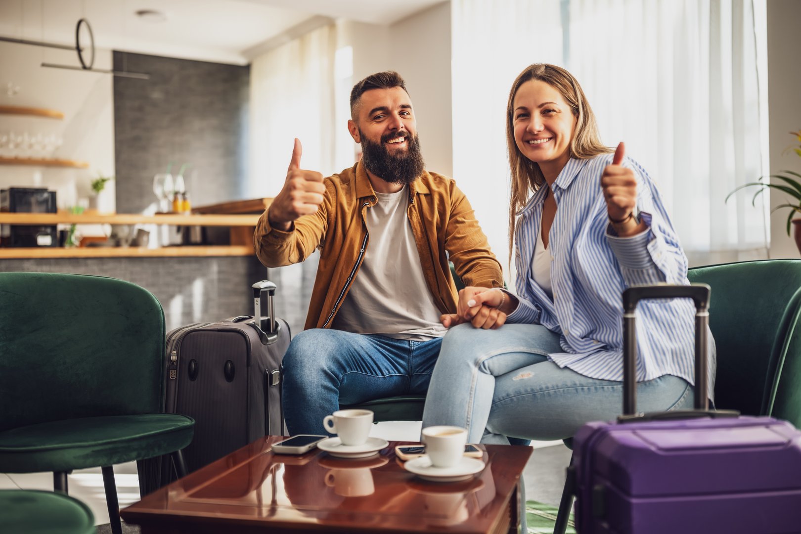 Married couple arrived in hotel. They are on vacation, sitting in lobby with suitcases, looking at camera, smiling and showing thumbs up.