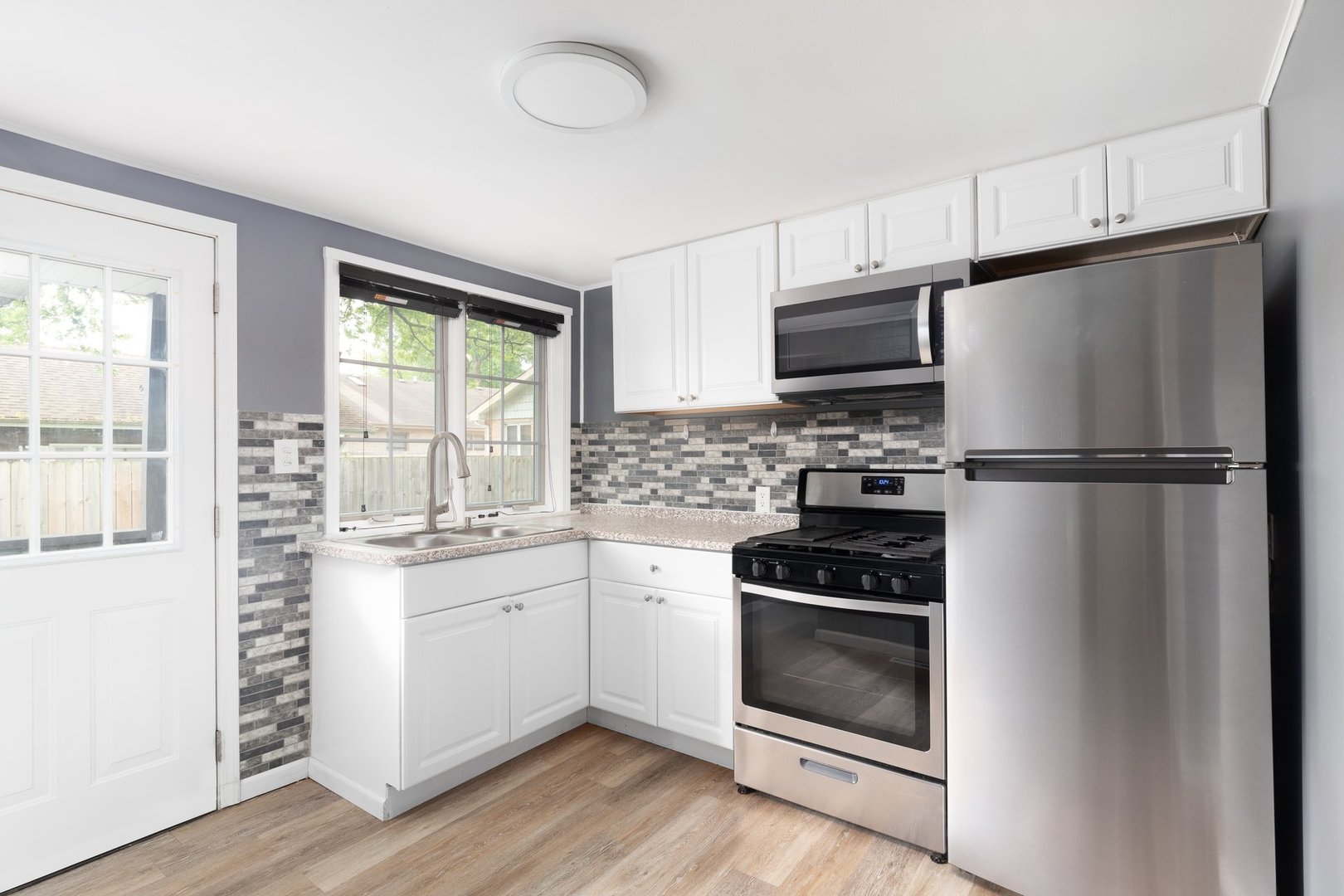 A small kitchen with white cabinets, stainless steel appliances, hardwood flooring, and a tiled backsplash.