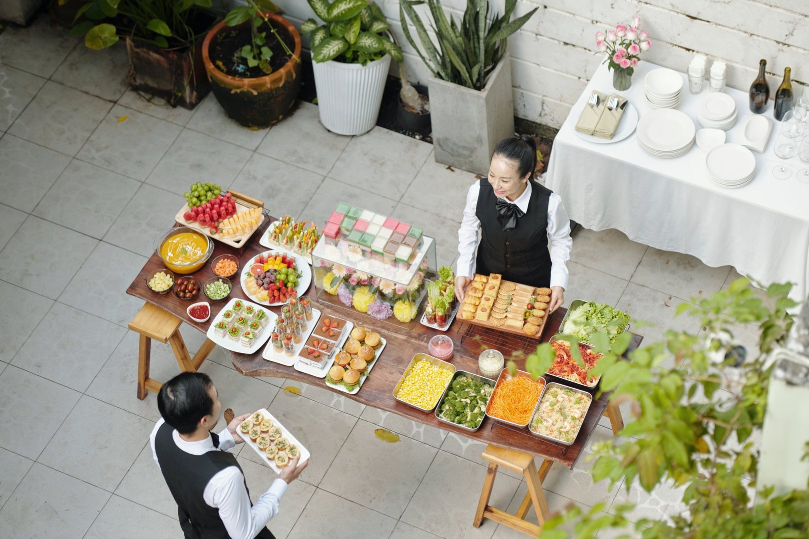 Overhead view of banquet setup with diverse foods and banquet attendants preparing dishes, featuring an array of colorful food items on a wooden table surrounded by plants