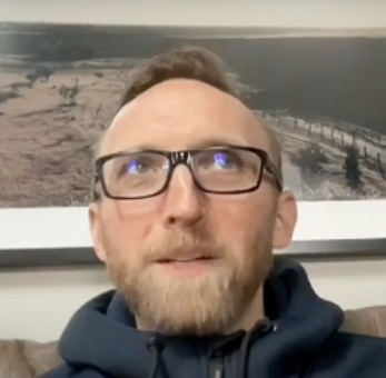 Man with glasses and beard wearing a dark hoodie, seated indoors with a landscape photo on the wall behind him.