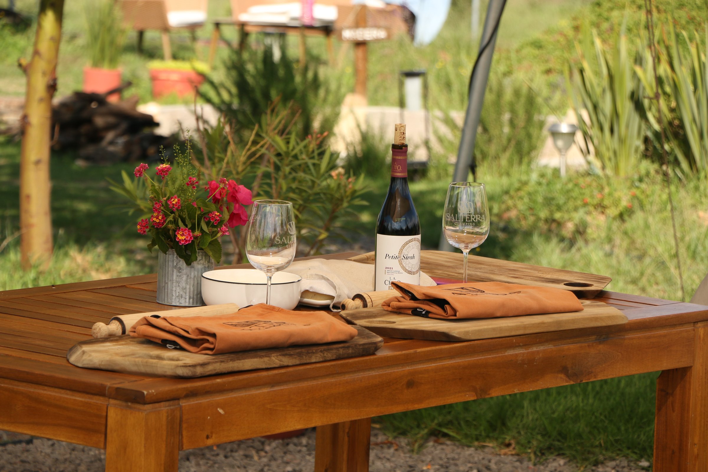 Outdoor table with wine bottle, two glasses, plates, rolled napkins, and flowers, set in a lush garden setting.