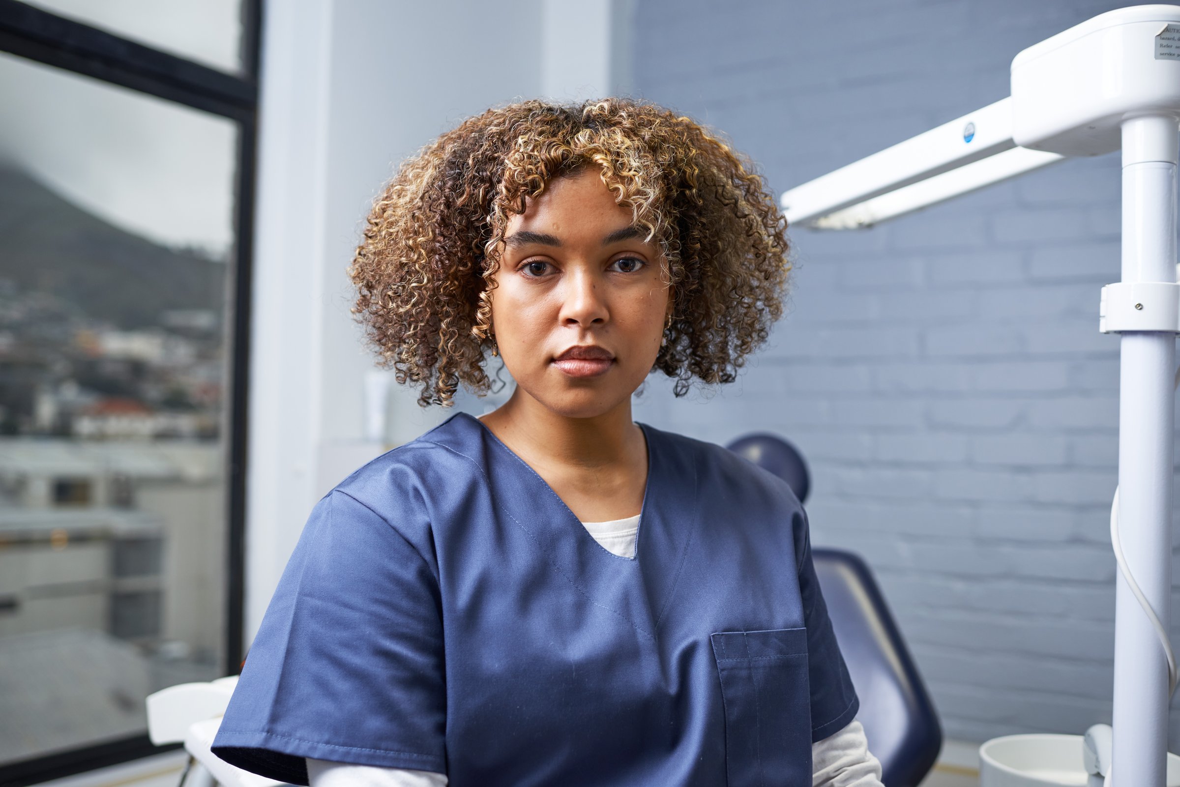 A healthcare professional dressed in scrubs poses in a dental clinic with natural light streaming through the window. The photo conveys professionalism and dedication within a healthcare environment.