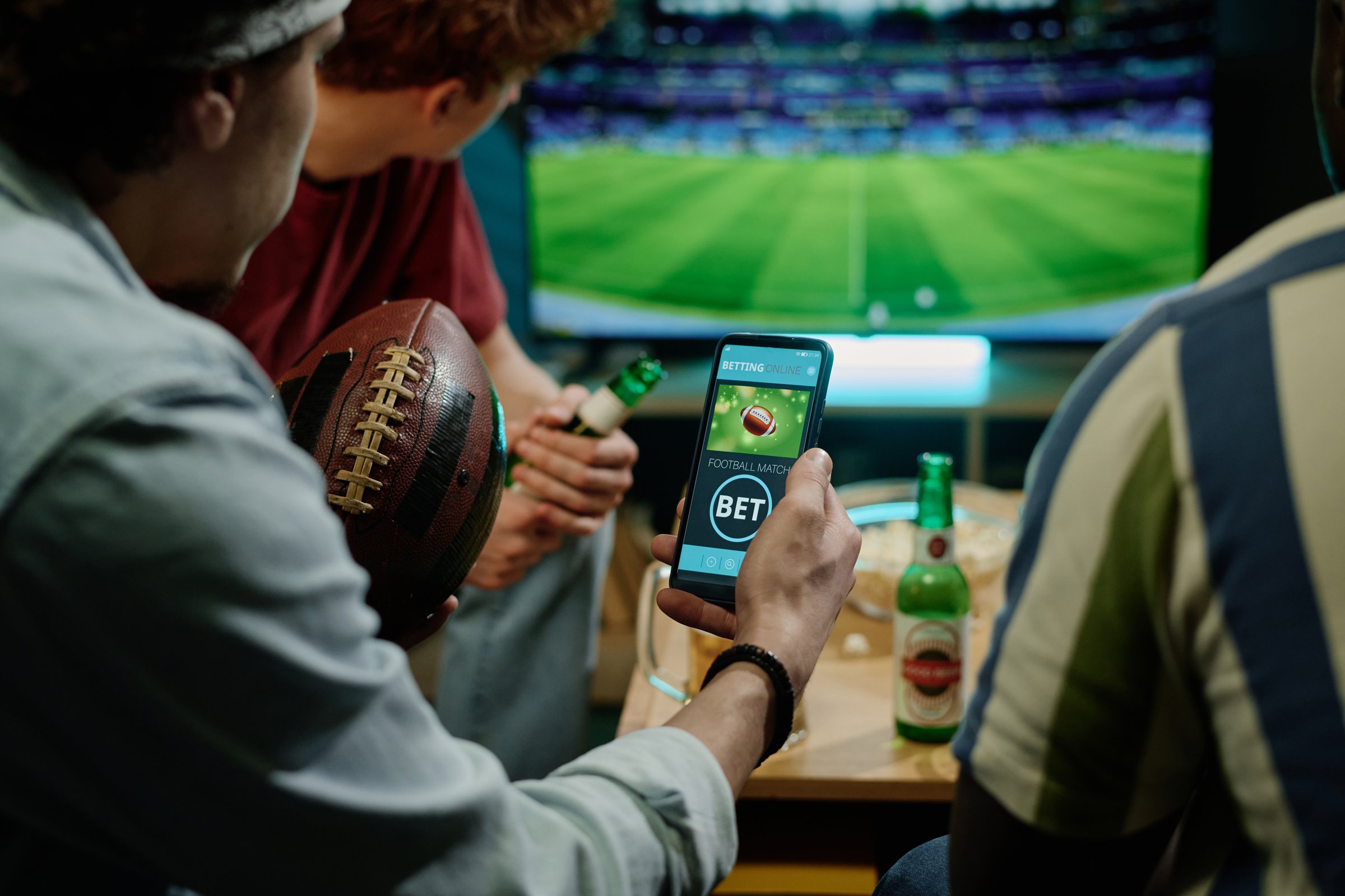 Crop shot of young male friends betting on team in app on smartphone while watching football match on TV