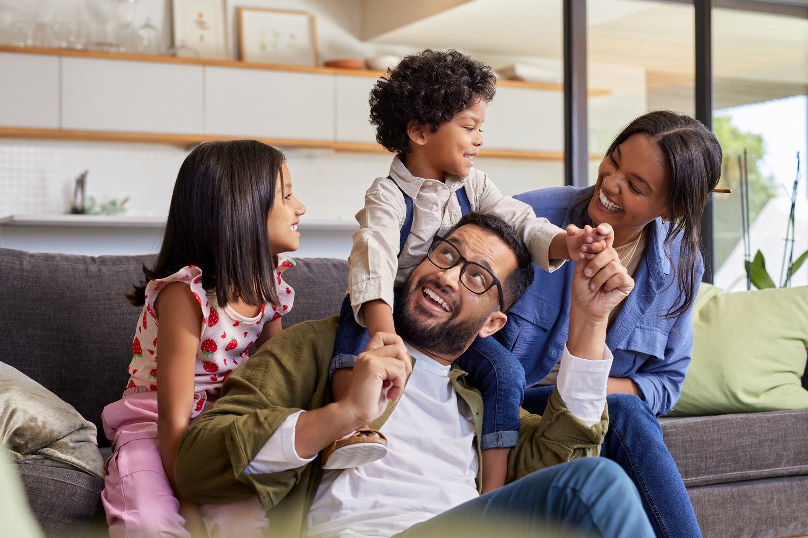 Happy multiethnic family with little son and daughter having fun at home. Happy middle eastern couple with son and daughter playing on sofa at home. Cute indian boy enjoying sitting on father shoulder while looking at mother.