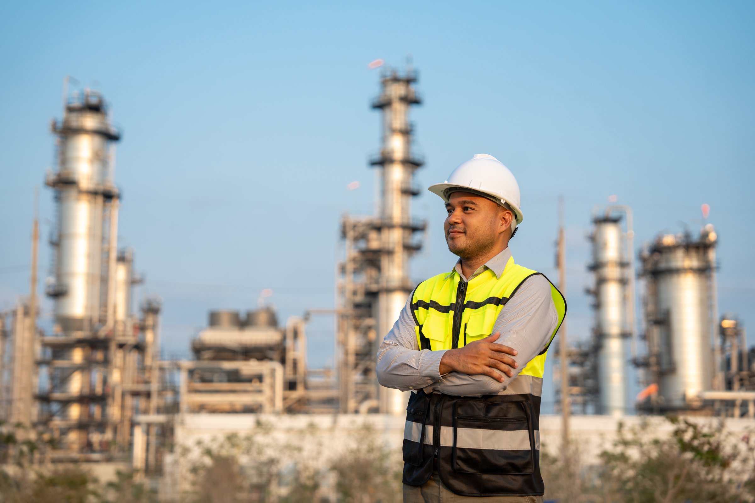 Asian engineer man with safety helmet standing front of oil refinery. Industry zone gas petrochemical. Factory oil storage tank and pipeline. Workers work in the refinery construction building