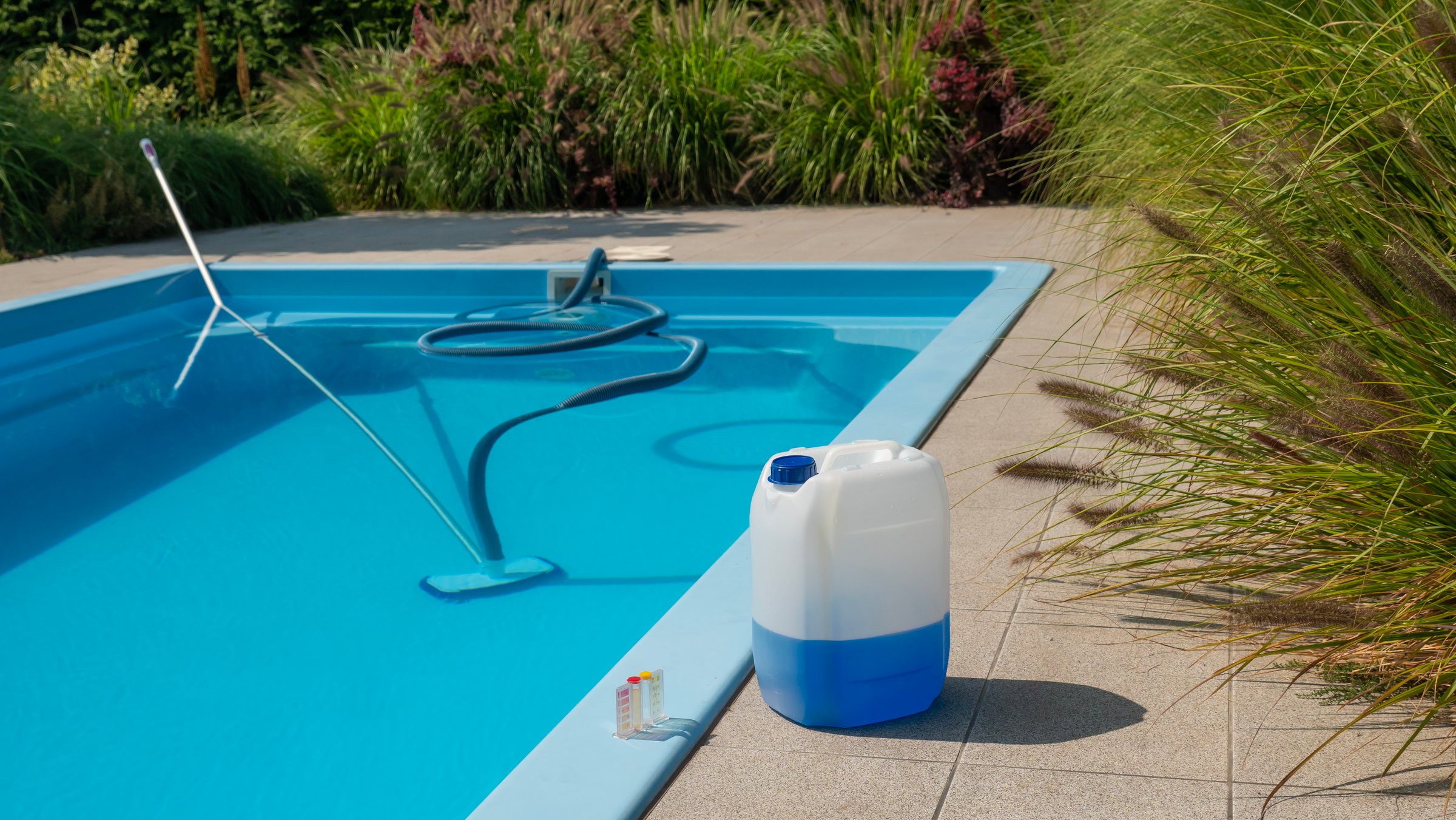 This image captures a pool cleaning hose and vacuum head submerged in a clear blue pool, with a large chemical container and water testing kit placed on the poolside. The lush greenery surrounding the pool creates a peaceful outdoor atmosphere, highlighting the essential tasks of maintaining water quality and ensuring a safe swimming environment.