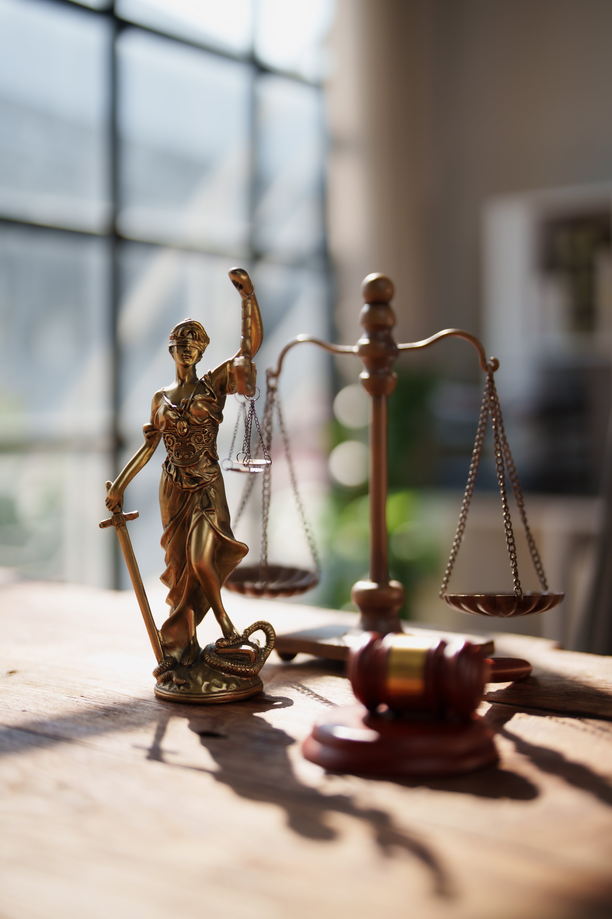 Lady Justice statue with scales and sword beside a gavel on a wooden legal desk, warm daylight highlighting the bronze figurine symbolizing fairness, balance, and authority